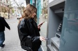 Young woman in leather jacket inserting a credit card to ATM outdoor, while looking on smartphone.